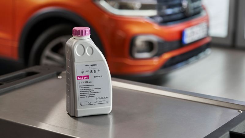 Flasche Original Volkswagen Kühlmittelzusatz G12evo auf einem Rollcontainer, im Hintergrund ein VW T-Cross