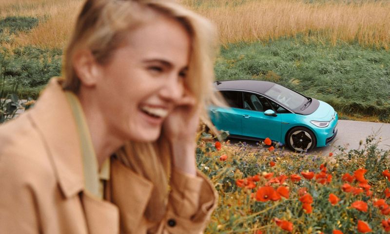 Eine Frau in einem schicken Trenchcoat lacht, im Hintergrund ist ein VW ID.3 zu sehen