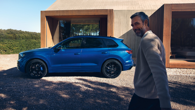 Un VW Touareg R eHybrid, vue en de profil devant une maison. Un homme se dirige vers lui en souriant.