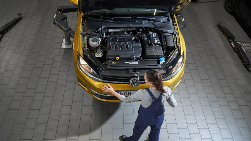 VW service employee standing in front of a VW car with open bonnet, focus on the engine compartment