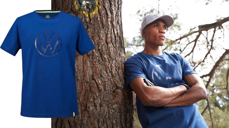 Ein Mann mit blauem T-Shirt mit VW Logo in 3D lehnt an einem Baum – Volkswagen Kollektion