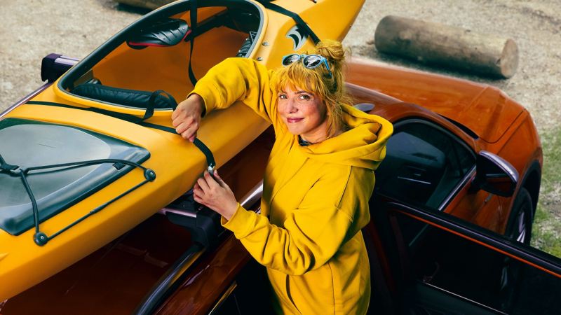 Eine Frau in Gelb an einem Volkswagen Auto greift nach ihrem Kajak – VW Transportprodukte