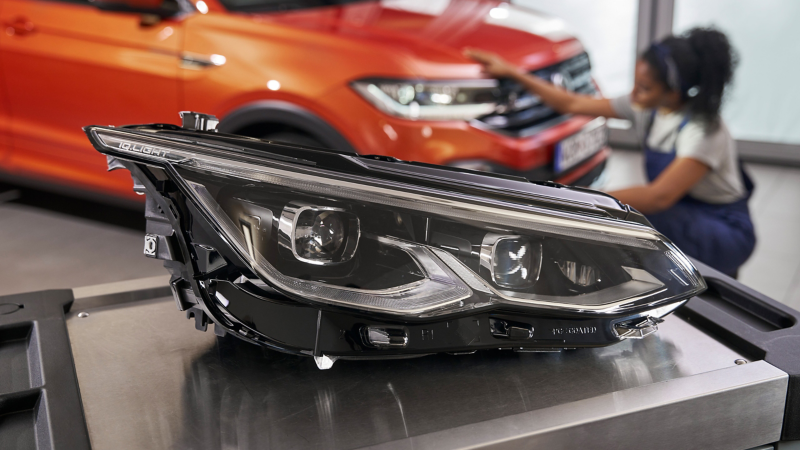  Un nouveau phare automobile est posé sur une surface métallique, avec une personne travaillant sur l'avant d'une voiture orange en arrière-plan flou.
