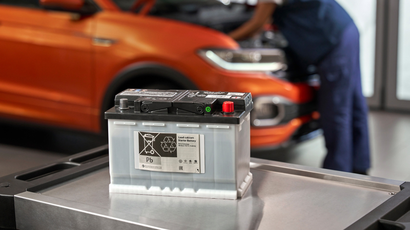 Volkswagen Genuine Lead-Calcium Starter Battery on a roller container, a VW T-Cross in the background