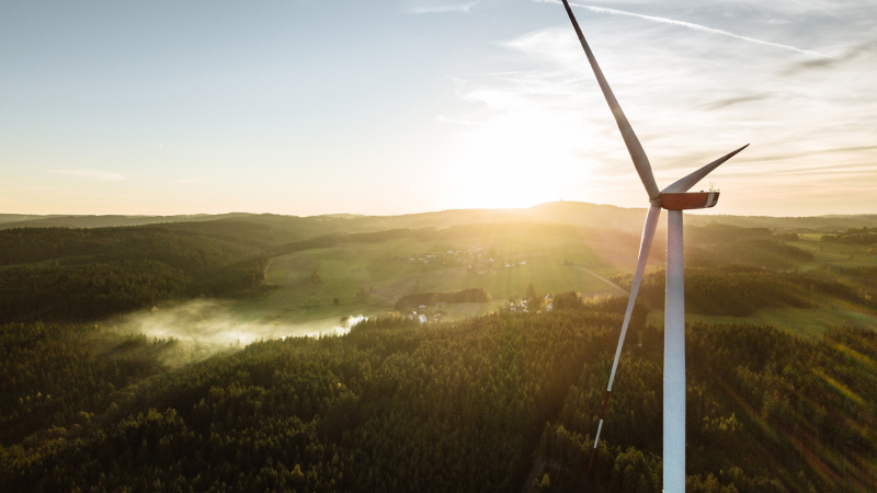Blick auf grüne Landschaft bei Sonnenuntergang, im Vordergrund Windrad – Naturstrom von VW