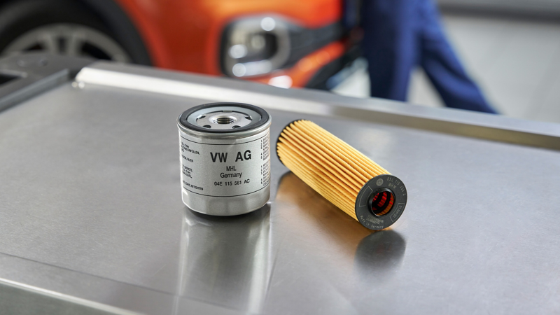  Un filtre à huile de type cartouche et un filtre à huile de type spin-on, marqués "VW AG", sont posés sur une surface métallique, avec une voiture orange et une personne floue en arrière-plan.