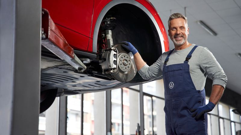 Ein VW Servicemitarbeiter neben einem VW Auto auf einer Hebebühne in der Werkstatt – Service für Elektroautos