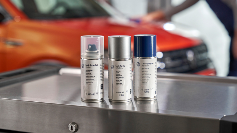 Trois bombes de peinture en aérosol de différentes couleurs (transparent, argent et bleu foncé) sont posées sur une surface métallique, avec l'avant flou d'une voiture orange en arrière-plan.