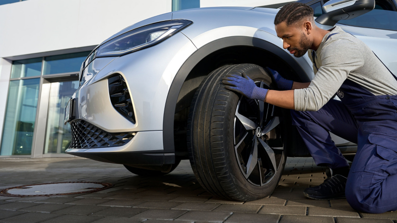  Un mécanicien agenouillé, portant des gants bleus, inspecte le pneu avant d'une voiture grise garée devant un bâtiment blanc.