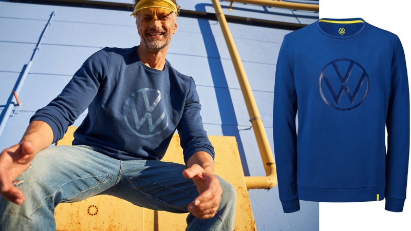 Mann mit blauem Sweatshirt mit VW Logo und einer gelben Schirmmütze in der Hocke – Volkswagen Kollektion