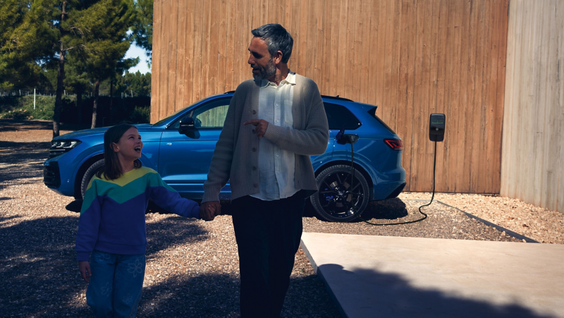 Un VW Touareg R eHybrid, vue de profil, est en charge devant une maison contemporaine. Au premier plan un homme et sa fille s'éoignent.