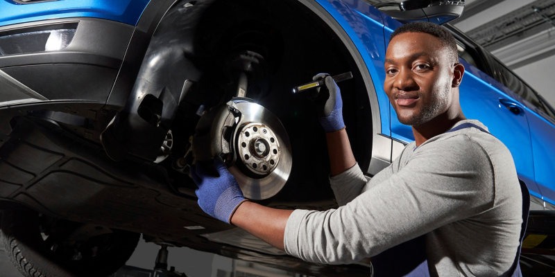 Un technicien de service souriant, portant des gants bleus, travaille sur le système de freinage d'une voiture bleue sur un élévateur dans un garage.