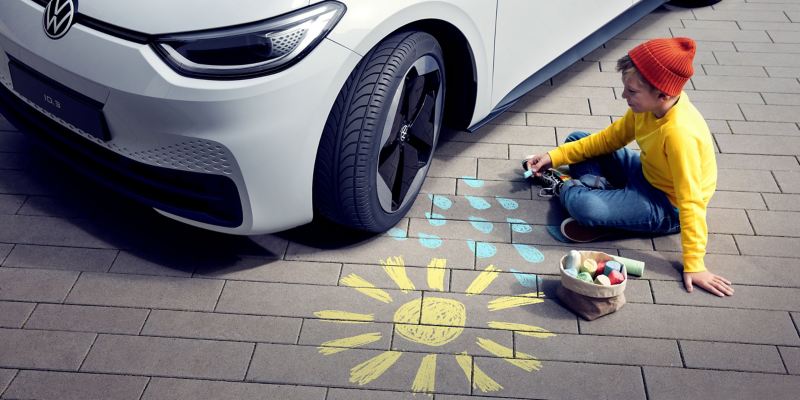 Et barn siddende foran en turkis VW ID.3 med Original +Dæk tegner et plus-mærke i en cirkel på vejen.