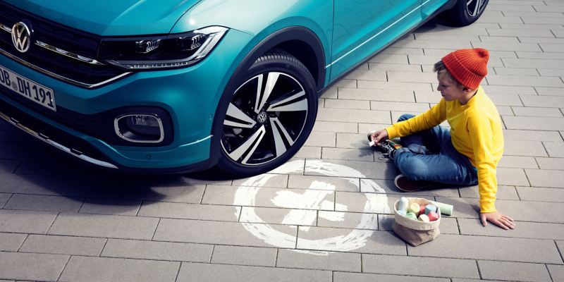Niño sentado frente a un VW T-Cross con neumáticos+, dibujando el símbolo más en un círculo en la calle