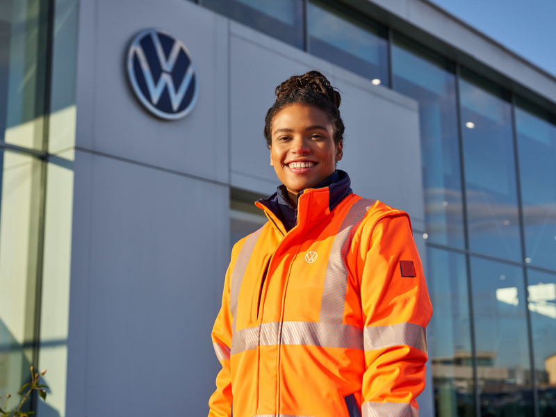 VW Servicemitarbeiterin in orangener Jacke vor Werkstatt