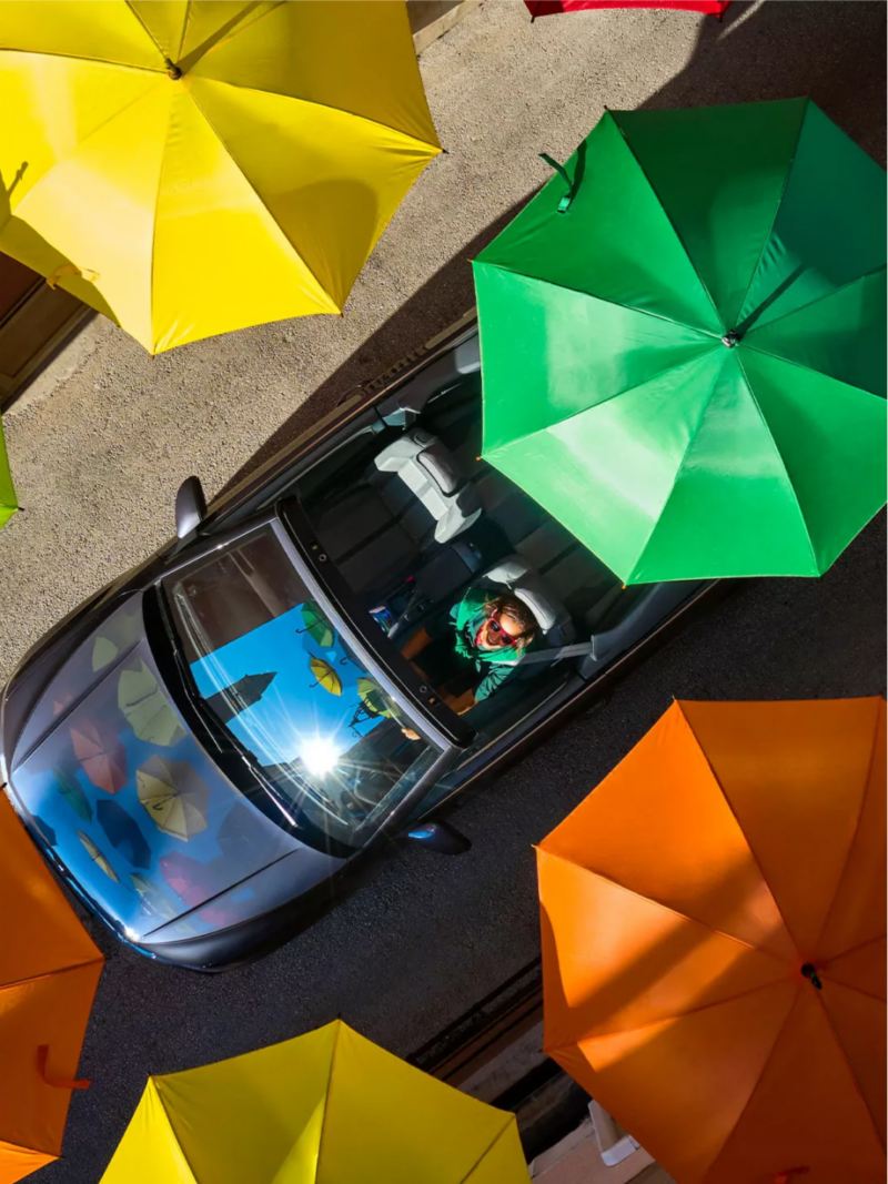 Convertible VW driving among colorful umbrellas.