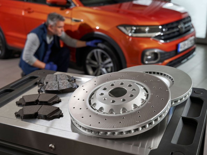 Disques de frein et plaquettes de frein d’origine Volkswagen sur une servante d’atelier, avec un VW T-Cross en arrière-plan