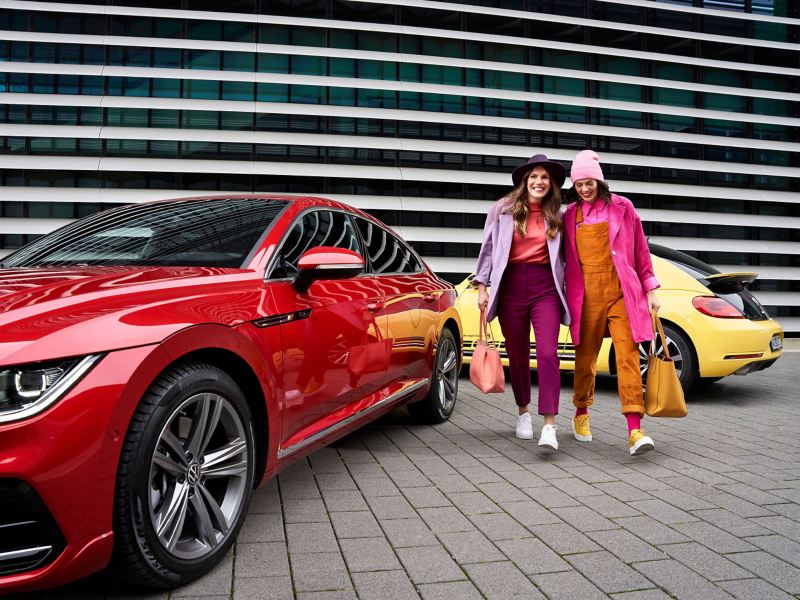 Zwei Frauen mit Einkaufstaschen neben ihren beiden Volkswagen – VW Aktionen und Angebote