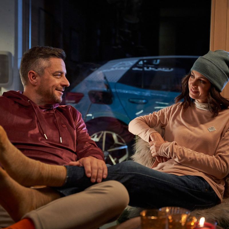 A couple is sitting comfortably in the living room, in the background an electric car is charging at the wall charging station