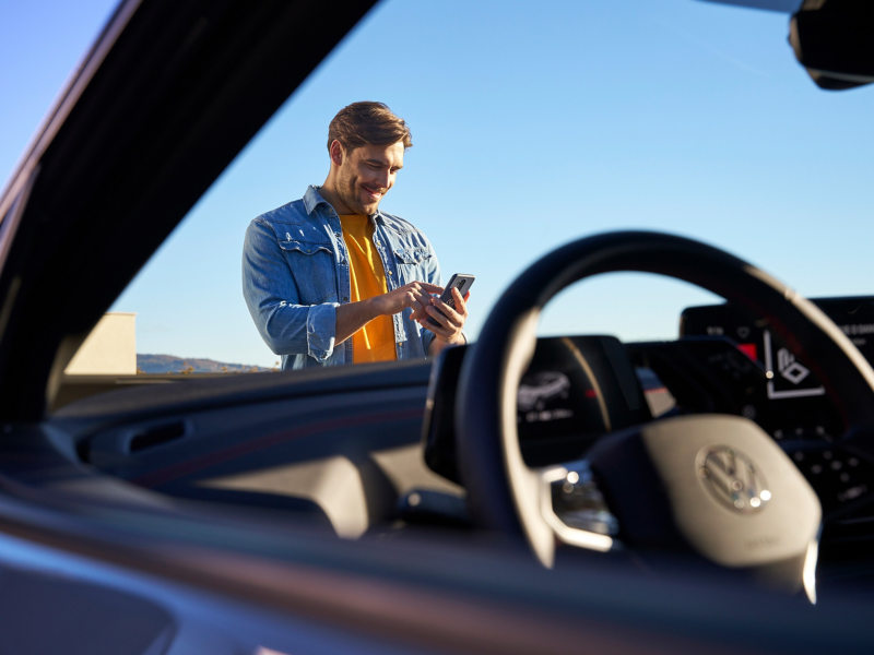  Un homme souriant en chemise en jean et t-shirt orange utilise son téléphone portable, vu de l'intérieur d'une Volkswagen avec le volant et le tableau de bord au premier plan. Le ciel est clair et bleu en arrière-plan.