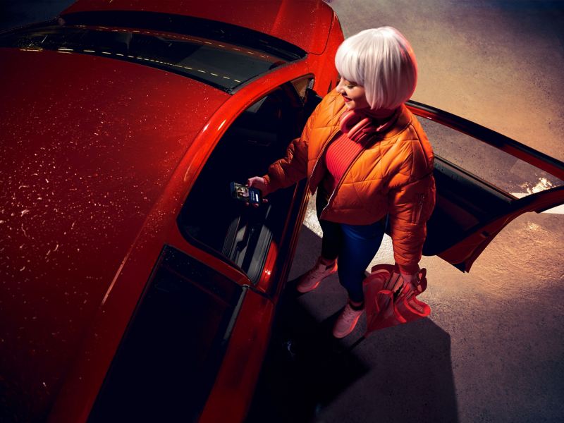 Une femme se tient à côté de la porte passager ouverte d’une VW Golf rouge et se connecte à la voiture via un smartphone