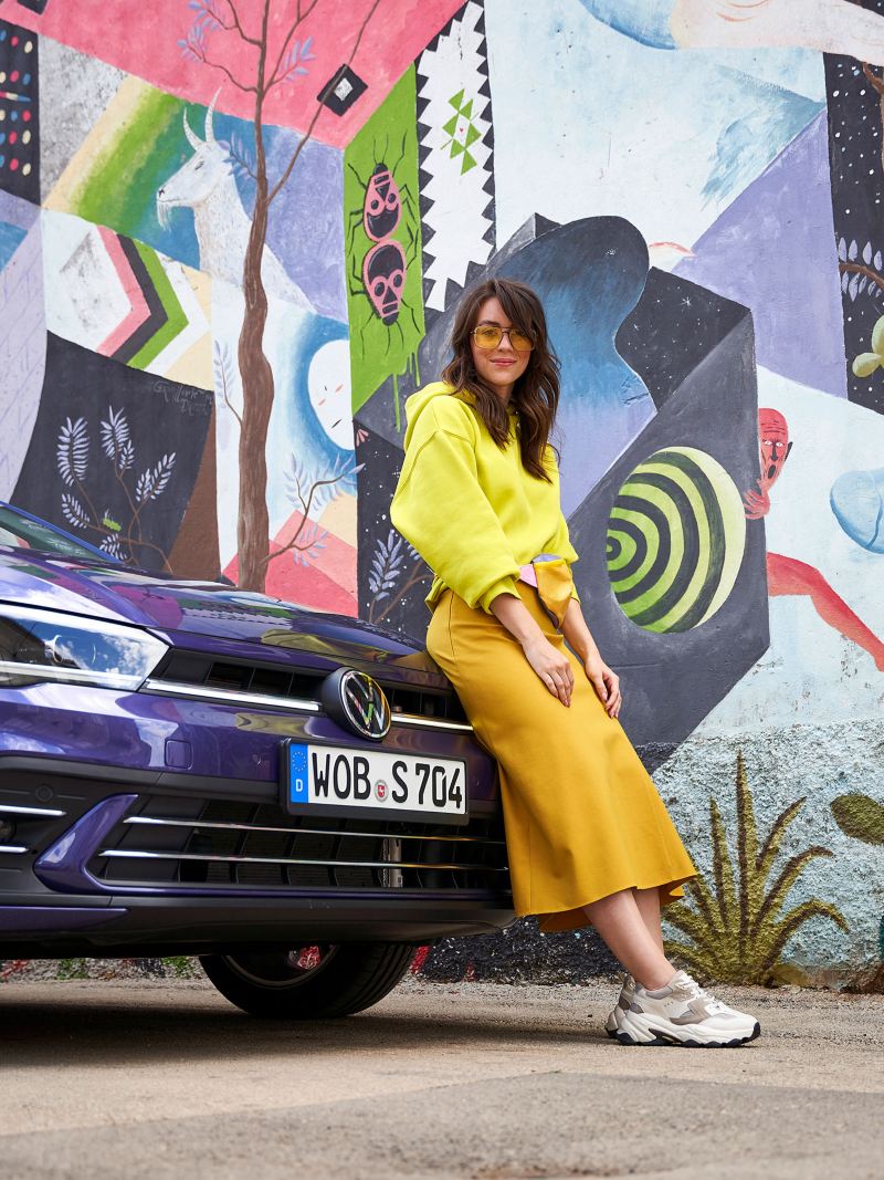 Eine junge Frau steht vor ihrem VW Auto vor einer bunten Wand