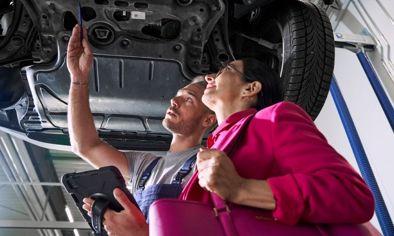 Un collaborateur d’atelier Volkswagen et une cliente regardent le dessous d’une voiture Volkswagen