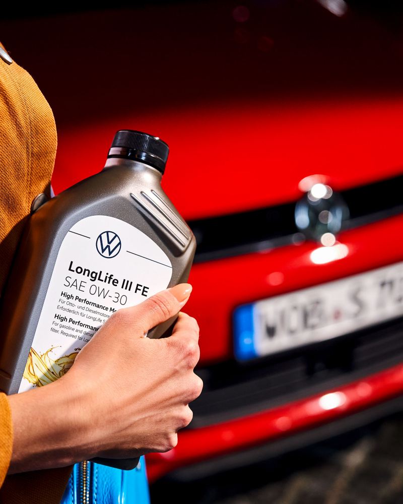 Μια γυναίκα κρατάει ένα μπουκάλι λαδιού κινητήρα στα χέρια της μπροστά από ένα Volkswagen
