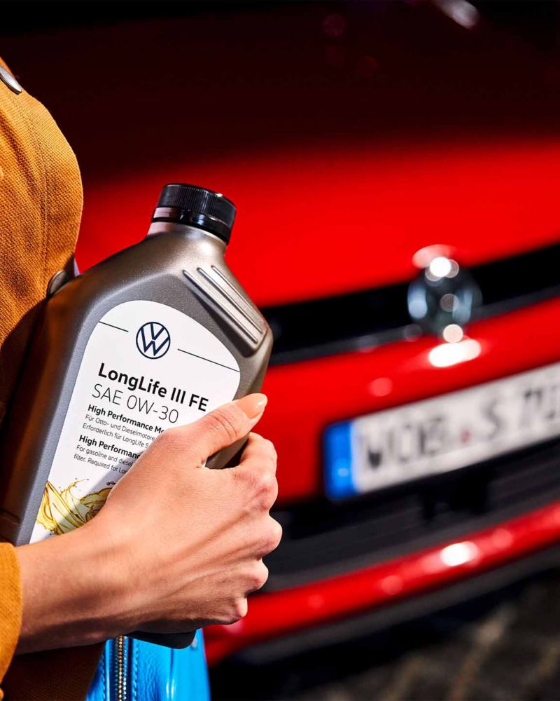 Vista di una mano femminile mentre tiene in  mano una confezione di olio per motore e sullo sfondo una vettura Volkswagen.
