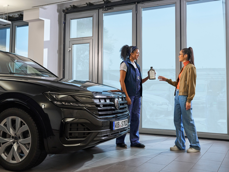 VW Servicemitarbeitender übergibt dem Kunden eine Flasche Motorenöl neben einem VW Touareg