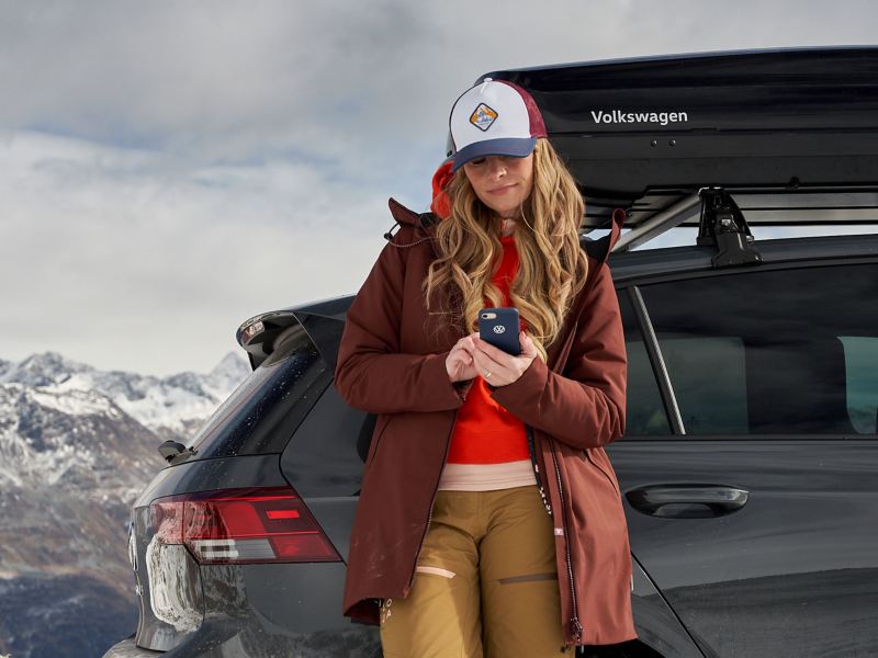 Une femme regarde son smartphone, porte une casquette de la Collection Heritage, et se tient devant sa voiture VW avec coffre de toit