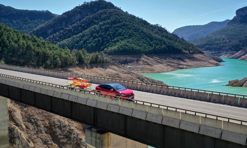Ένα VW ID.5 GTX που μεταφέρει με τρέιλερ ένα καγιάκ οδηγεί πάνω σε μια γέφυρα με μια λίμνη στο βάθος – λύσεις μεταφοράς από τα Αξεσουάρ VW