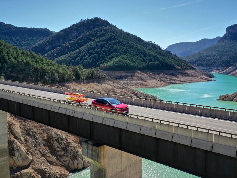 Ένα VW ID.5 GTX που μεταφέρει με τρέιλερ ένα καγιάκ οδηγεί πάνω σε μια γέφυρα με μια λίμνη στο βάθος – λύσεις μεταφοράς από τα Αξεσουάρ VW
