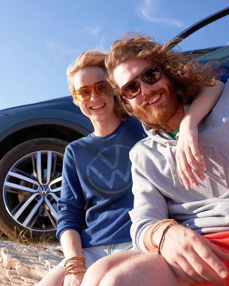 Eine Frau und ein Mann in stylischer Kleidung sitzen vor Ihrem VW Auto – VW Fanartikel