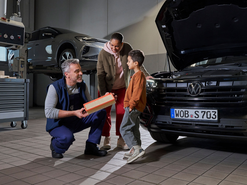 pracownik serwisu pokazujący chłopcu części eksploatacyjne Volkswagena, obok chłopca stoi matka, w tle serwisowane w warsztacie samochody