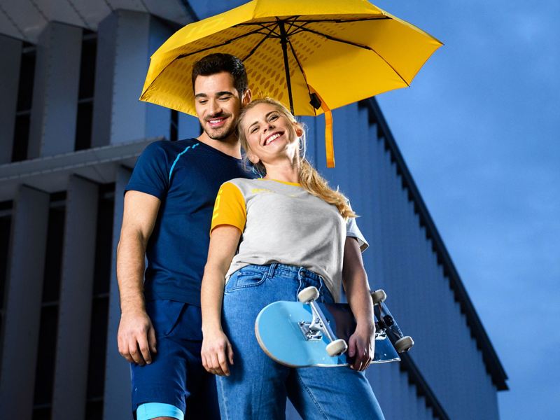 Ein lachendes Pärchen mit moderner ID. Kleidung unter einem gelben Regenschirm – VW Merchandise