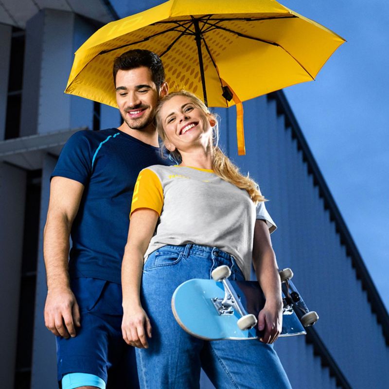Ein lachendes Pärchen mit moderner ID. Kleidung unter einem gelben Regenschirm – VW Merchandise