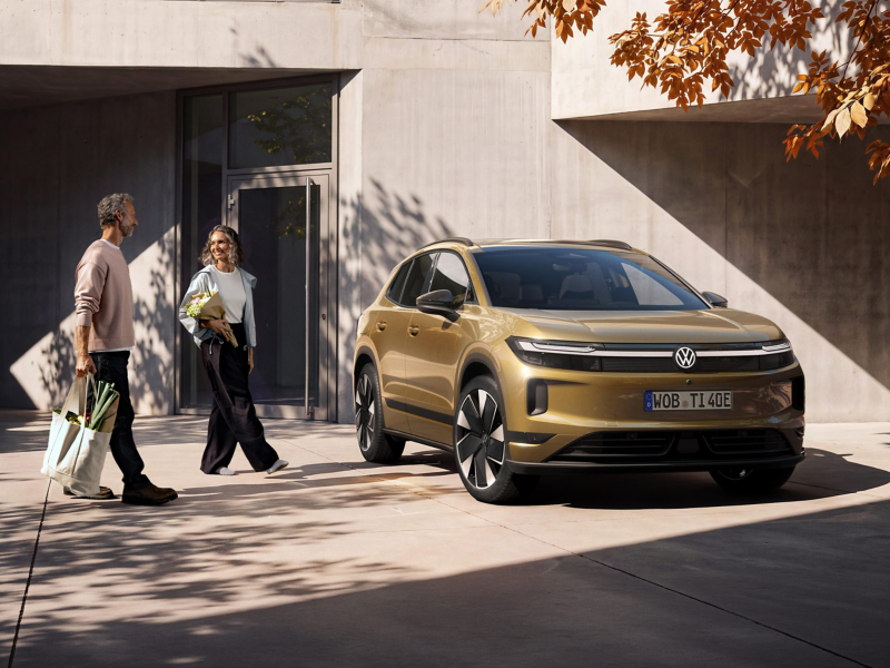 Ein Mann und eine Frau gehen mit Einkäufen beladen auf einen oliv-bronzefarbenen VW ID. Tiguan zu, der vor einem modernen Gebäude parkt.