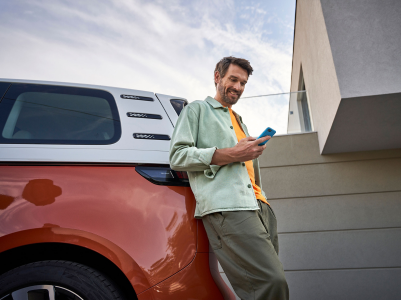 Un homme souriant se tient appuyé contre un Volkswagen ID. Buzz bicolore orange et blanc et connecte son smartphone à son véhicule avec App Connect