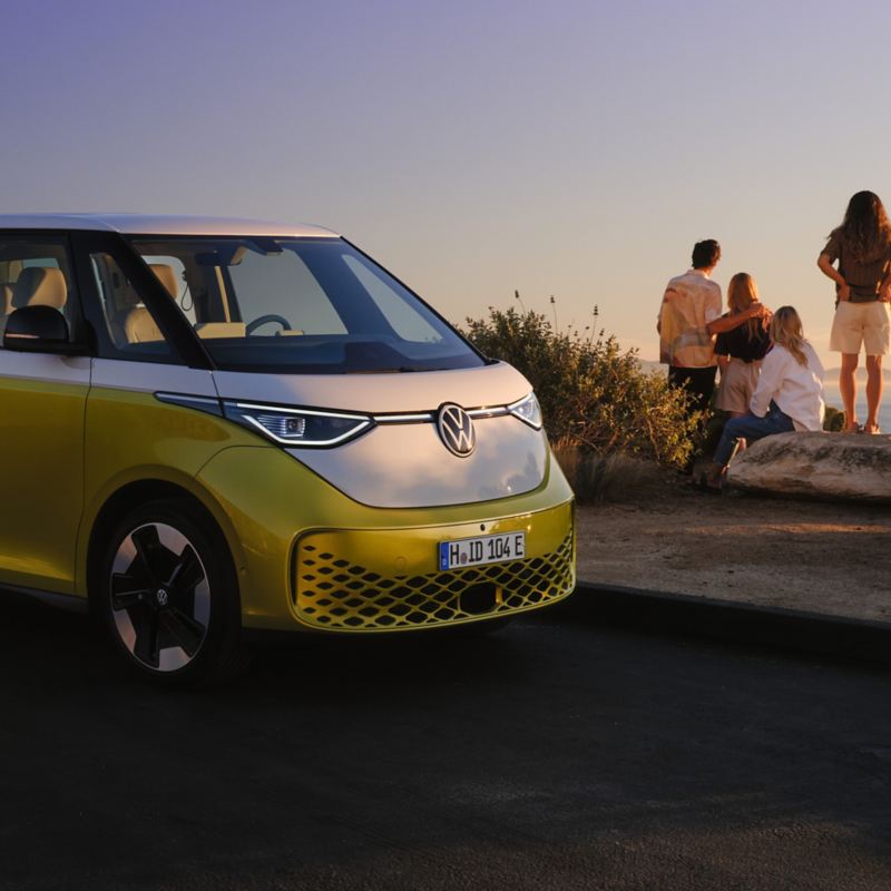 Ein gelb-weißer VW ID. Buzz am Rand einer Strandpromenade, im Hintergrund eine Familie mit zwei Kindern am Meer