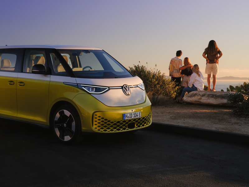 Ein gelb-weißer VW ID. Buzz am Rand einer Strandpromenade, im Hintergrund eine Familie mit zwei Kindern am Meer