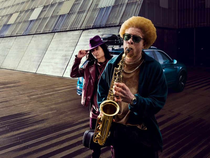 A man with a saxophone and a man with a hat in front of a VW car