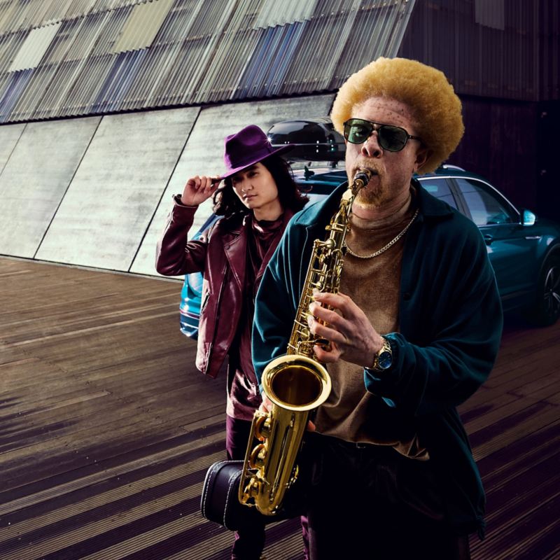 A man with a saxophone and a man with a hat in front of a VW car