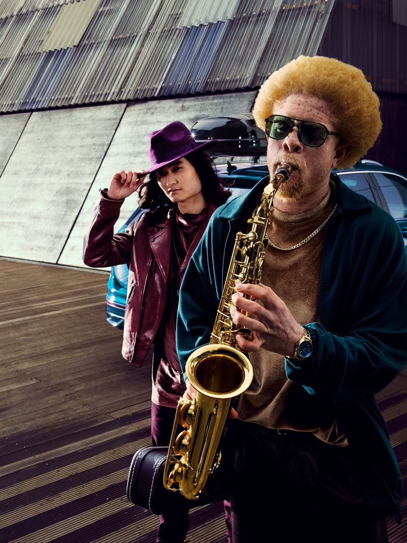 Un homme avec un saxophone et un homme avec un chapeau devant une VW