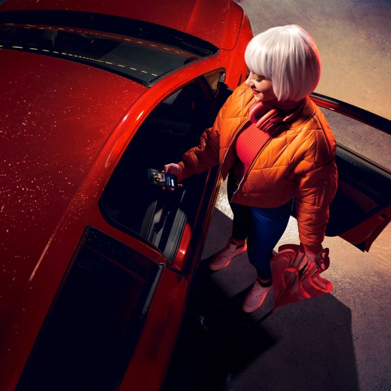 Eine Frau steht neben der offenen Beifahrertür eines roten VW Golf und verbindet sich per Smartphone mit dem Auto