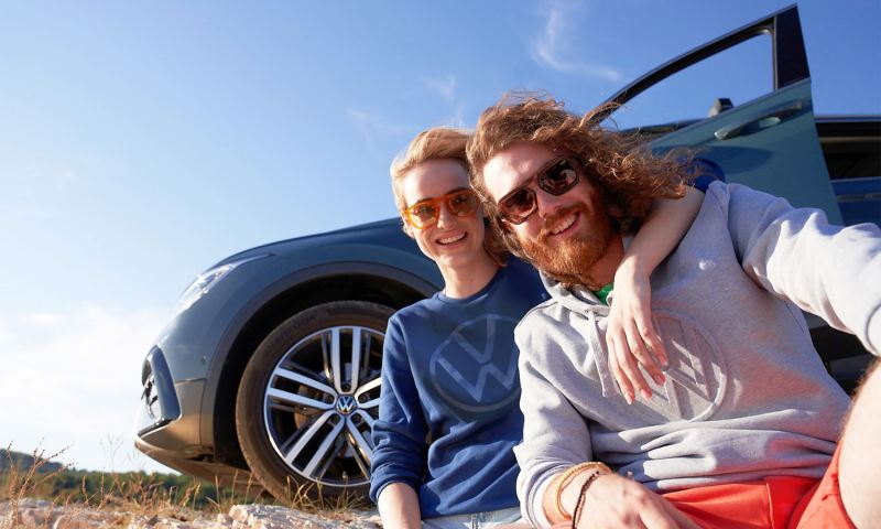 Μια γυναίκα και ένας άνδρας με κομψά ρούχα κάθονται μπροστά από το VW τους – προϊόντα VW Lifestyle