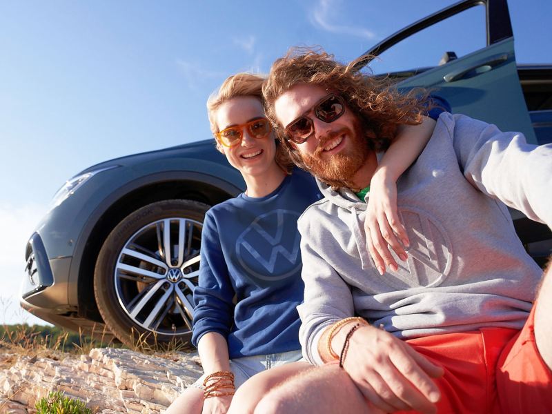 A woman and man in stylish clothes sitting in front of their VW car – VW Lifestyle products
