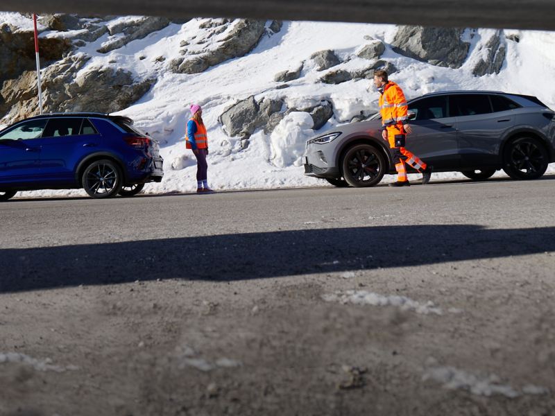 Ein Servicemitarbeiter vom Volkswagen Notdienst (Pannen- und Unfallhilfe) kommt einer Kundin zur Hilfe – im Hintergrund verschneite Berge und zwei VW Autos