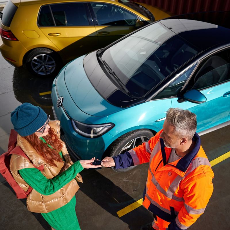 Eine Kundin überreicht einem VW Servicemitarbeiter ihre Autoschlüssel, im Hintergrund stehen zwei Volkswagen