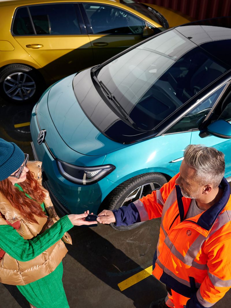 Eine Kundin überreicht einem VW Servicemitarbeiter ihre Autoschlüssel, im Hintergrund stehen zwei Volkswagen